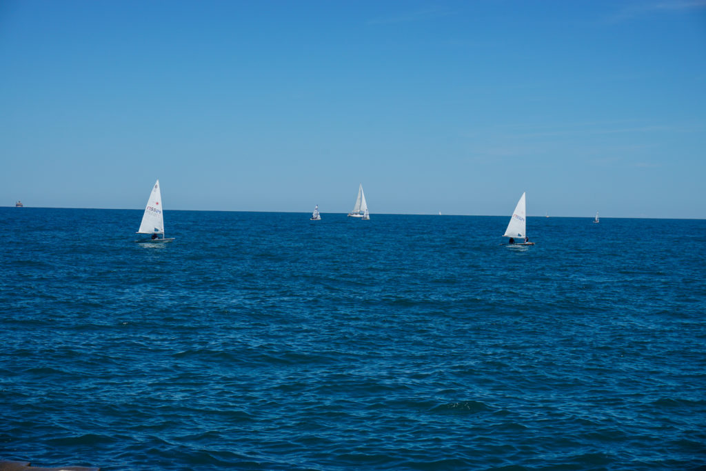 This photograph by Justin is dominated by two fields of blue, the sky which has no clouds, just a soft gradation as it reaches from the top of the image to the horizon, and the lake which is a much deeper blue and has the indication of gentle waves. On the lake are four sail boats, each with a white triangular sail which breaks across the horizon line and into the sky.