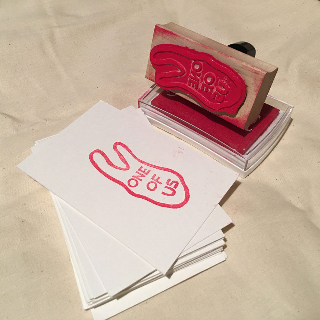 This artwork consists of a few separate elements: a stamp, an inkpad, and blank notecards. The hand-held stamp has a short black handle attached to a rectangular wood base. The stamp image is a hand with two fingers and on the palm are the words “one of us” The stamp pad is a rectangular plastic container holding a red felt pad. The blank note cards are small white rectangles intended to be stamped on and either taken or shared with the gallery.